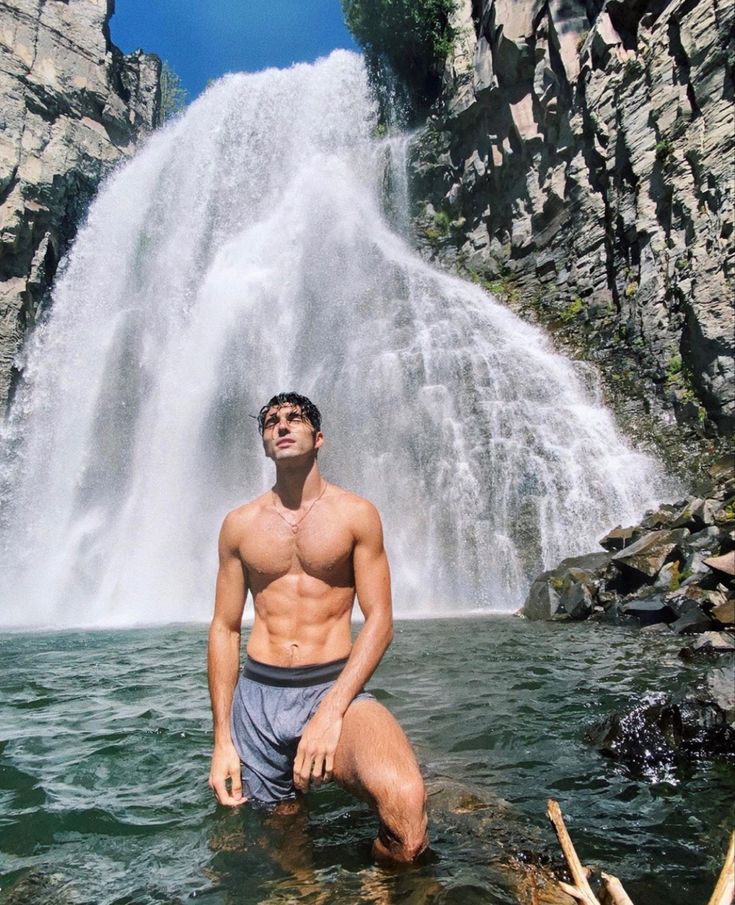 a shirtless man standing in front of a waterfall