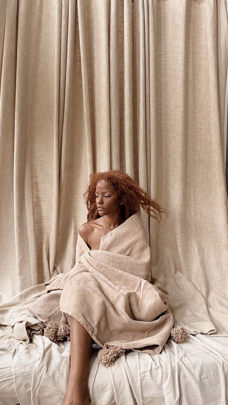 a woman wrapped in a blanket sitting on top of a bed next to a curtain