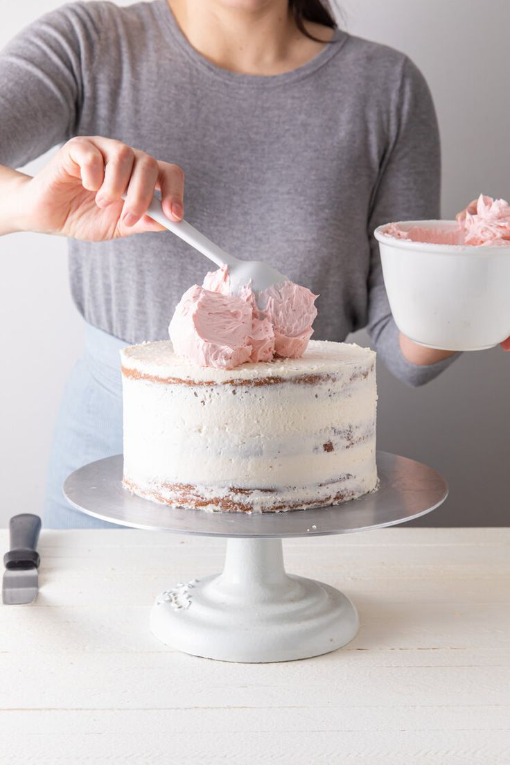 a woman is decorating a cake with pink frosting and a white bowl in front of her