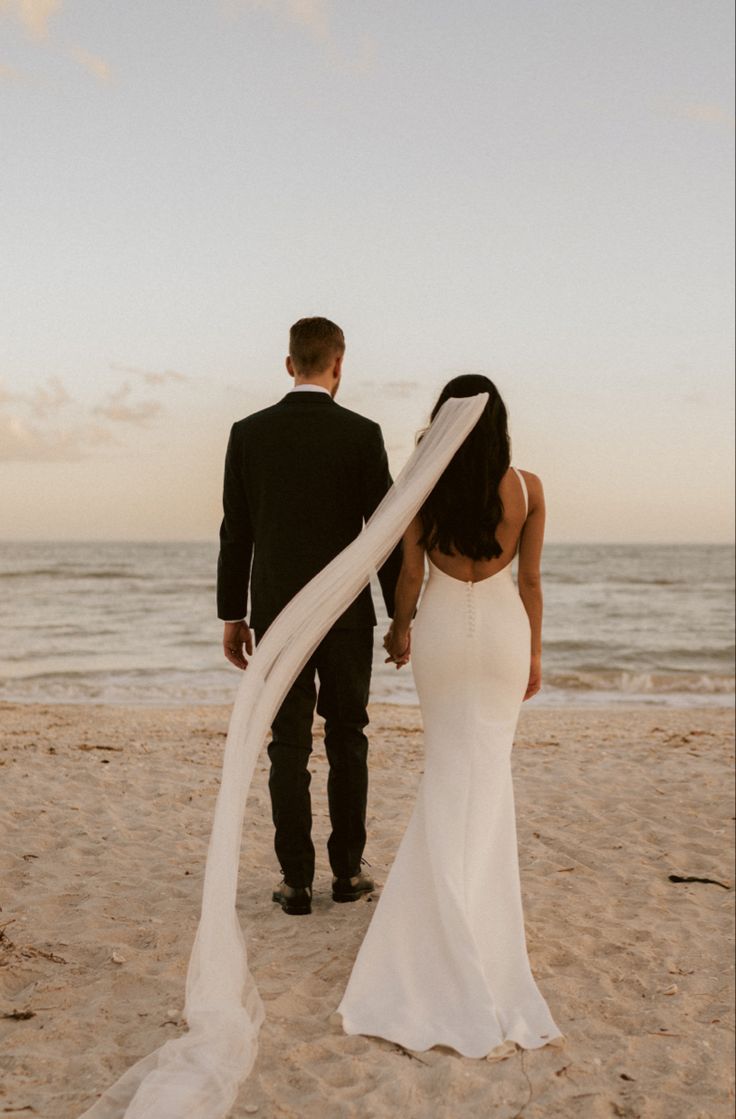 a bride and groom walking on the beach with their veil blowing in the wind as the sun sets