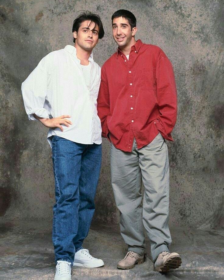 two men standing next to each other in front of a cement wall with their hands on their hips