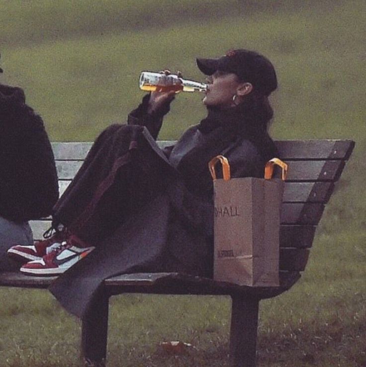 two people sitting on a park bench drinking beer and eating food from paper bags in front of them