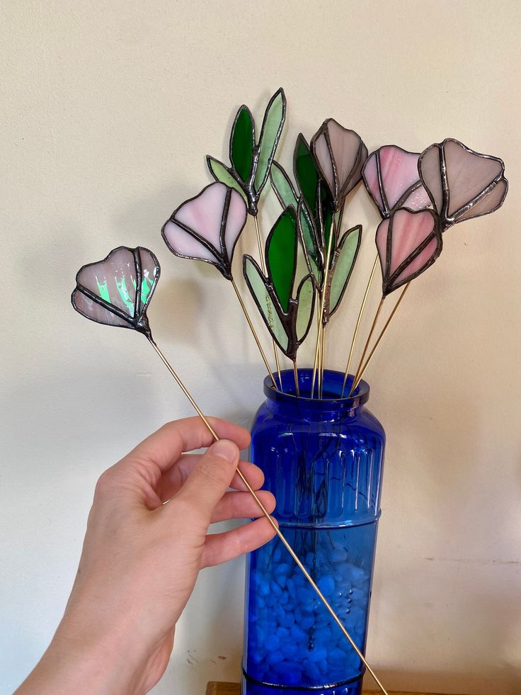 a blue vase filled with pink and green flowers next to a hand holding a stick