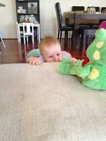 a baby laying on the floor next to a green stuffed animal and a table with chairs