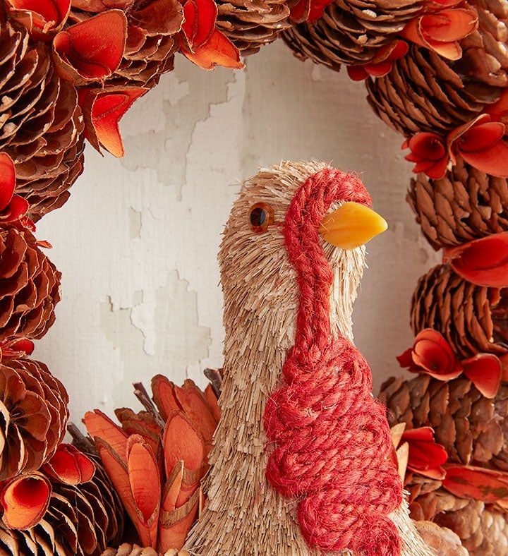 a chicken made out of pine cones and yarn sits in front of a wreath with red flowers