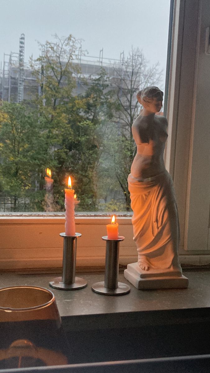 two candles are lit in front of a window with a statue on the windowsill