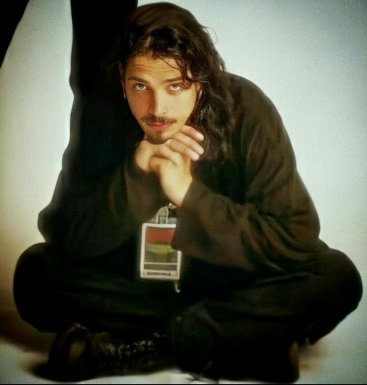 a man with long hair sitting on the ground next to a white wall and holding his hand under his chin