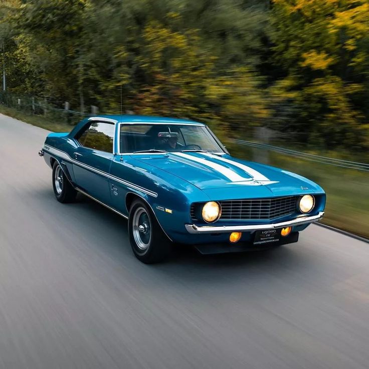 a blue muscle car driving down the road with trees in the background