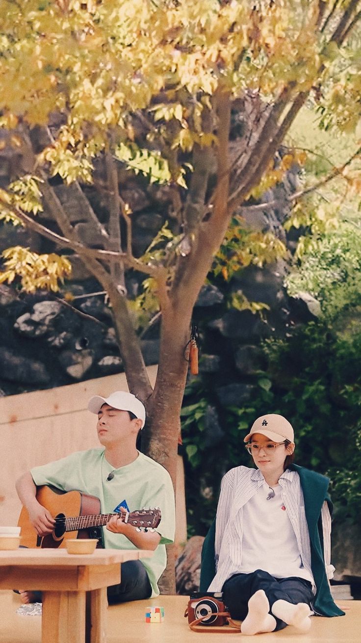 two people sitting at a picnic table playing guitars and drinking wine in front of a tree