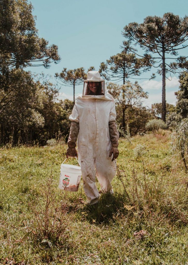 a man in a bee suit holding a bucket and walking through the grass with trees behind him