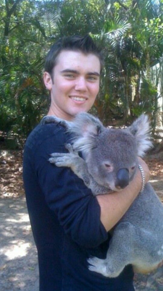 a man holding a koala bear in his arms and smiling at the camera with trees behind him