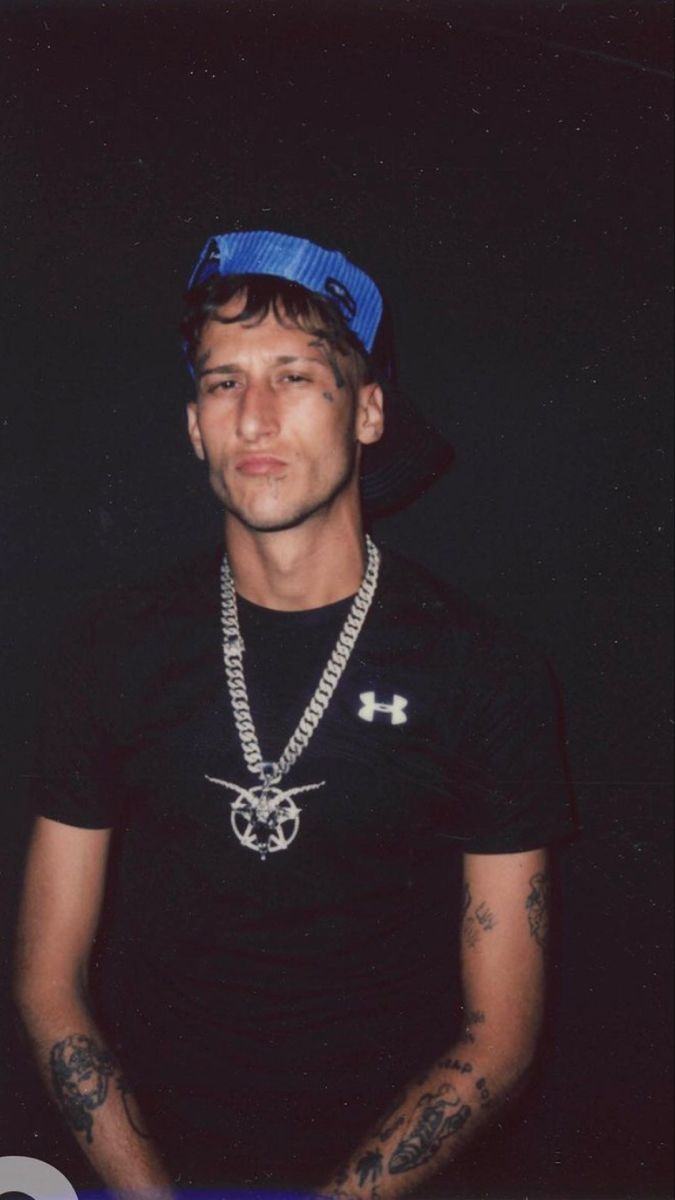 a young man wearing a black shirt and blue hat with tattoos on his arms, standing in front of a dark background