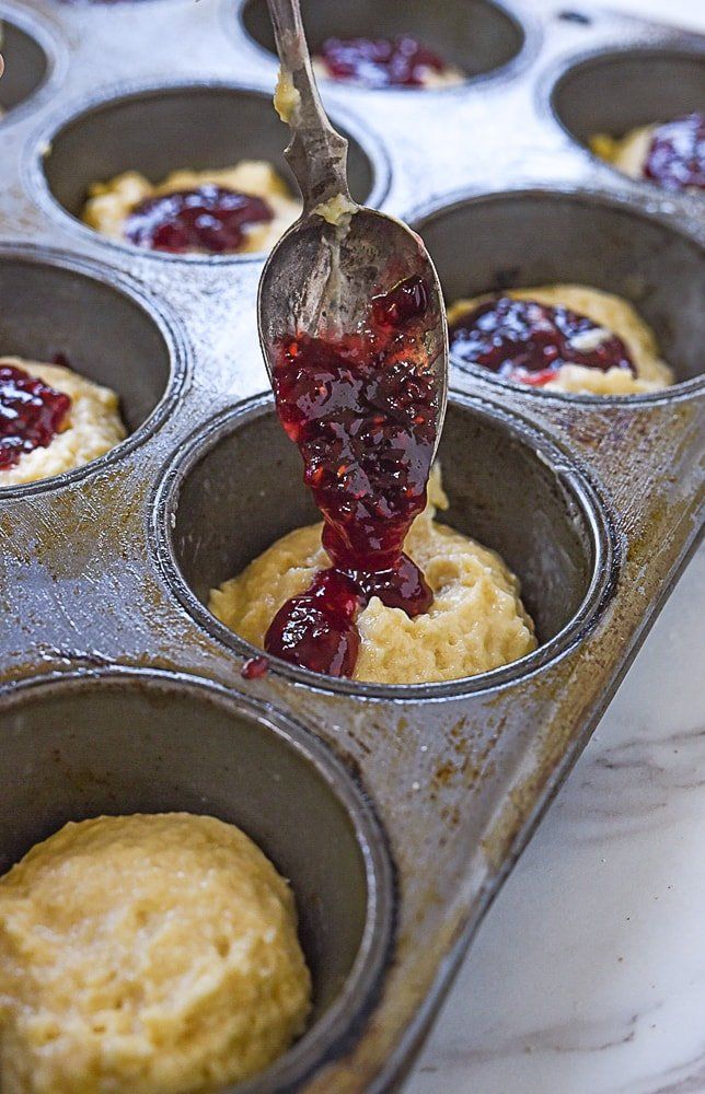 a muffin tin filled with cupcakes and jam being spooned into it