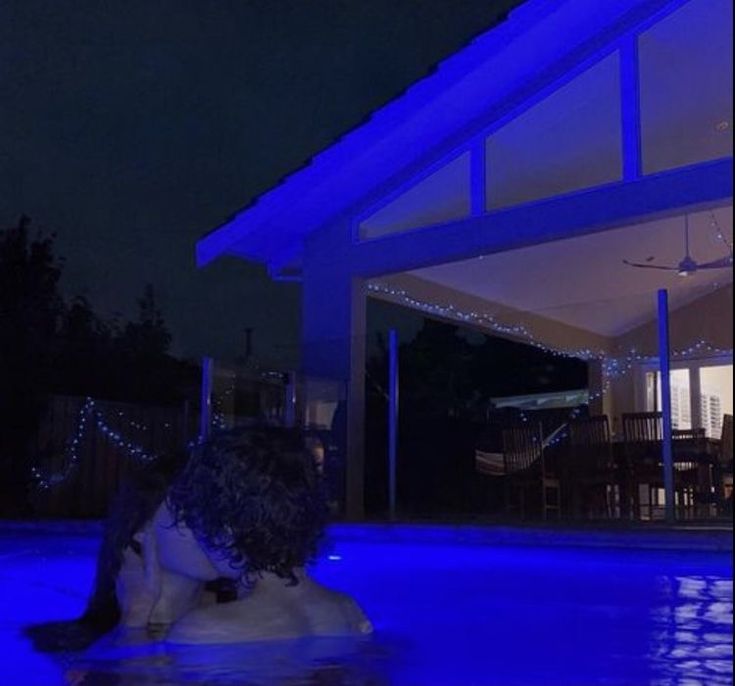 a woman sitting on the edge of a swimming pool at night with blue lights around her