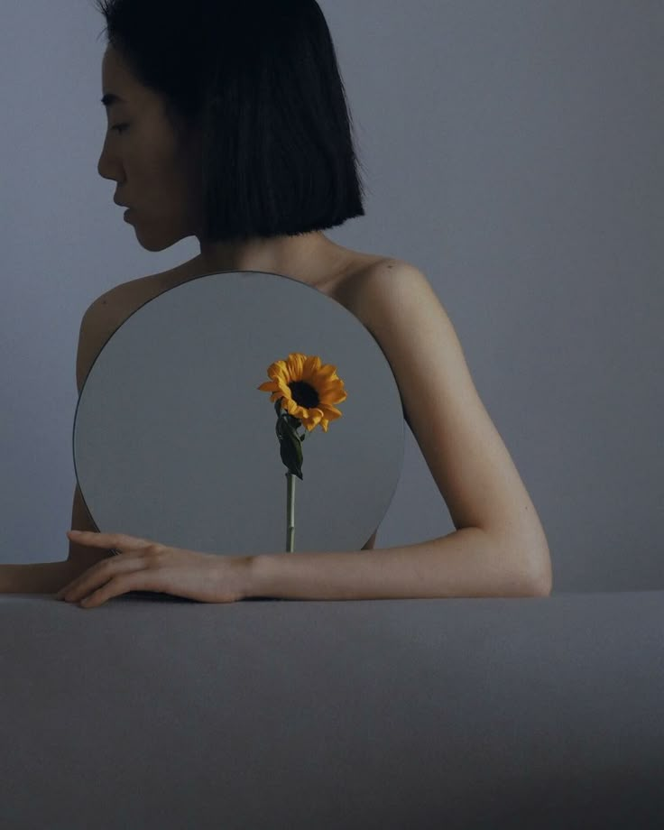 a woman holding a mirror with a sunflower in front of her face on the table