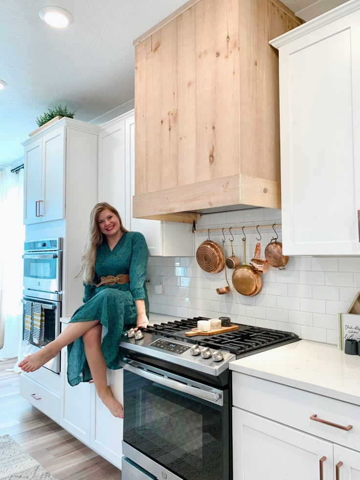 a woman sitting on top of a stove in a kitchen next to white cupboards