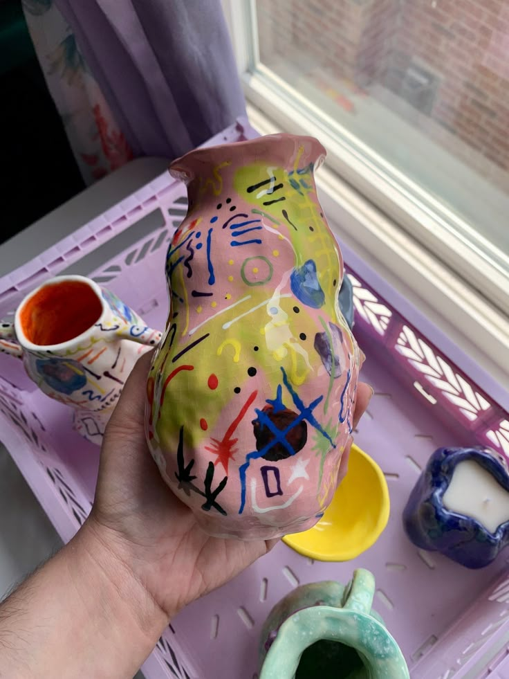 a hand holding a ceramic vase with colorful designs on it and two cups next to it