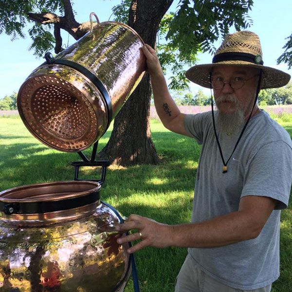 a man wearing a hat and holding a large copper pot in front of a tree