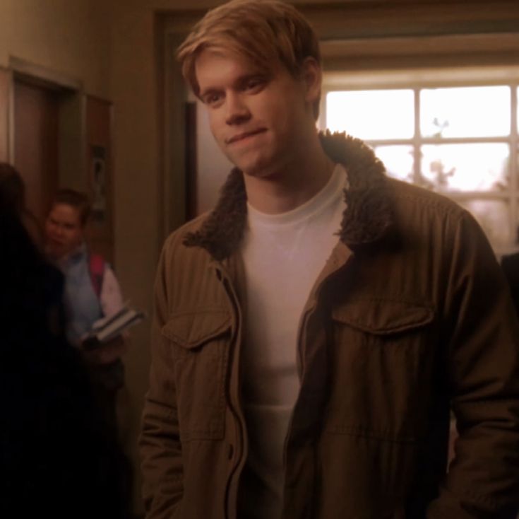 a young man in a brown jacket is looking at the camera while people are standing around him