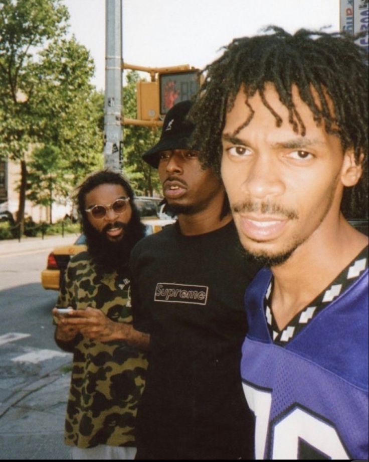 a man with dreadlocks standing in front of other men on the street and looking at his cell phone