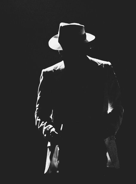 a man wearing a hat and coat in the dark with his hand on his hip