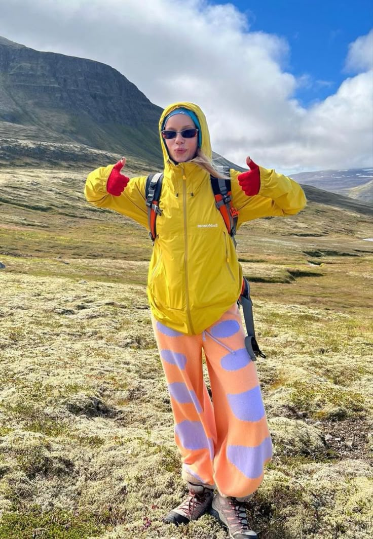 a woman in yellow jacket standing on top of a grass covered field with mountains in the background