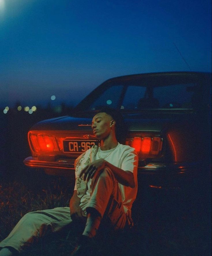 a man sitting on the ground in front of a car at night with its lights on