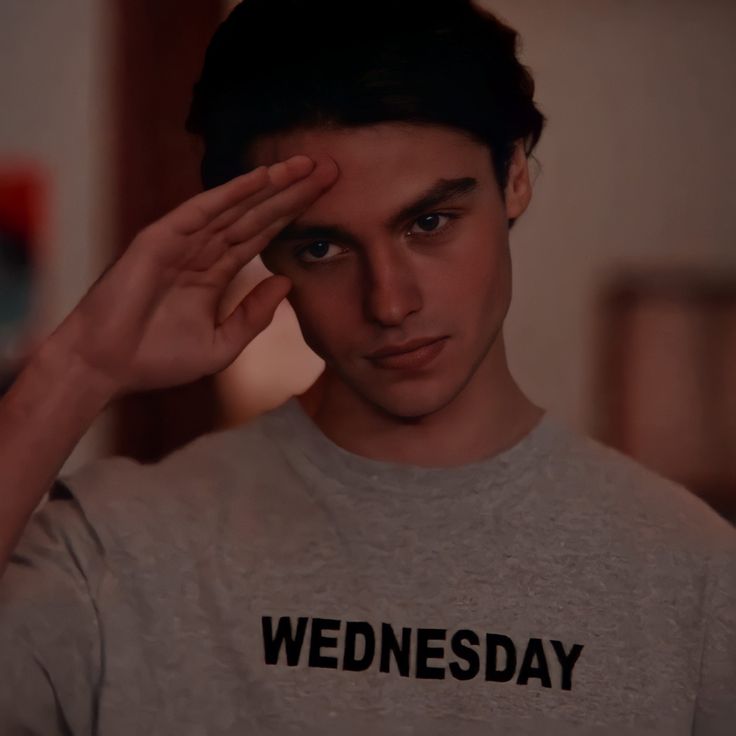 a young man holding his hand to his head and looking at the camera while wearing a wednesday t - shirt