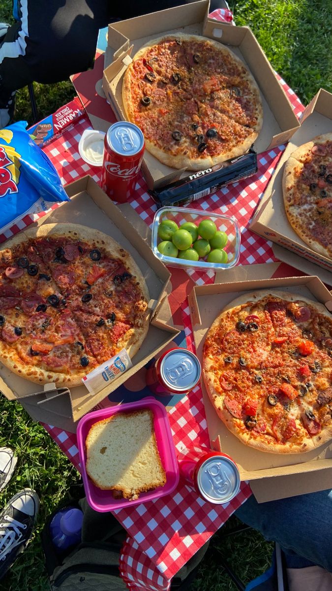 several pizzas and drinks are sitting on a picnic table with red checkered cloth