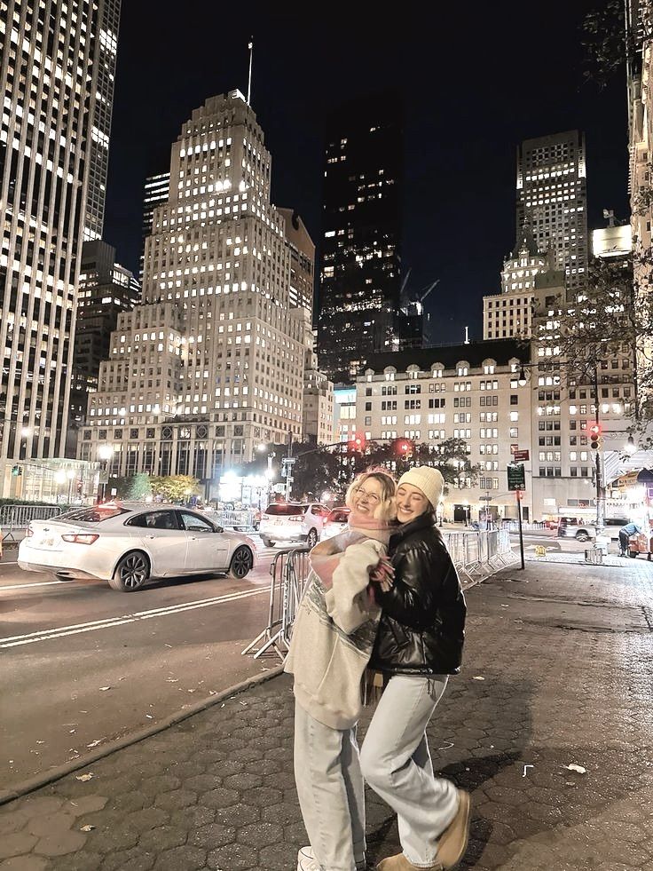 two people standing in the middle of a city at night, one is hugging the other