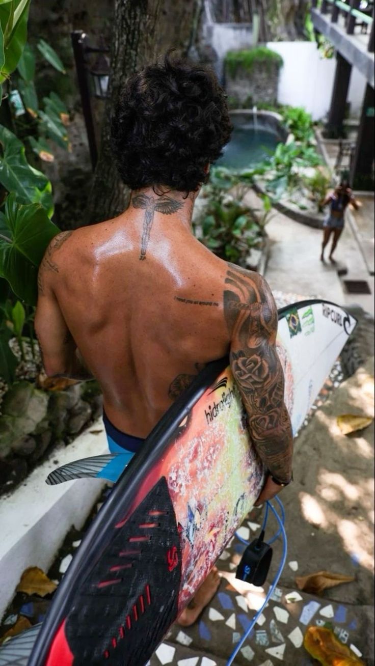 a man holding a surfboard while standing next to a tree and building with graffiti on it