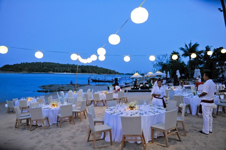 people are sitting at tables on the beach with white linens and lights strung over them