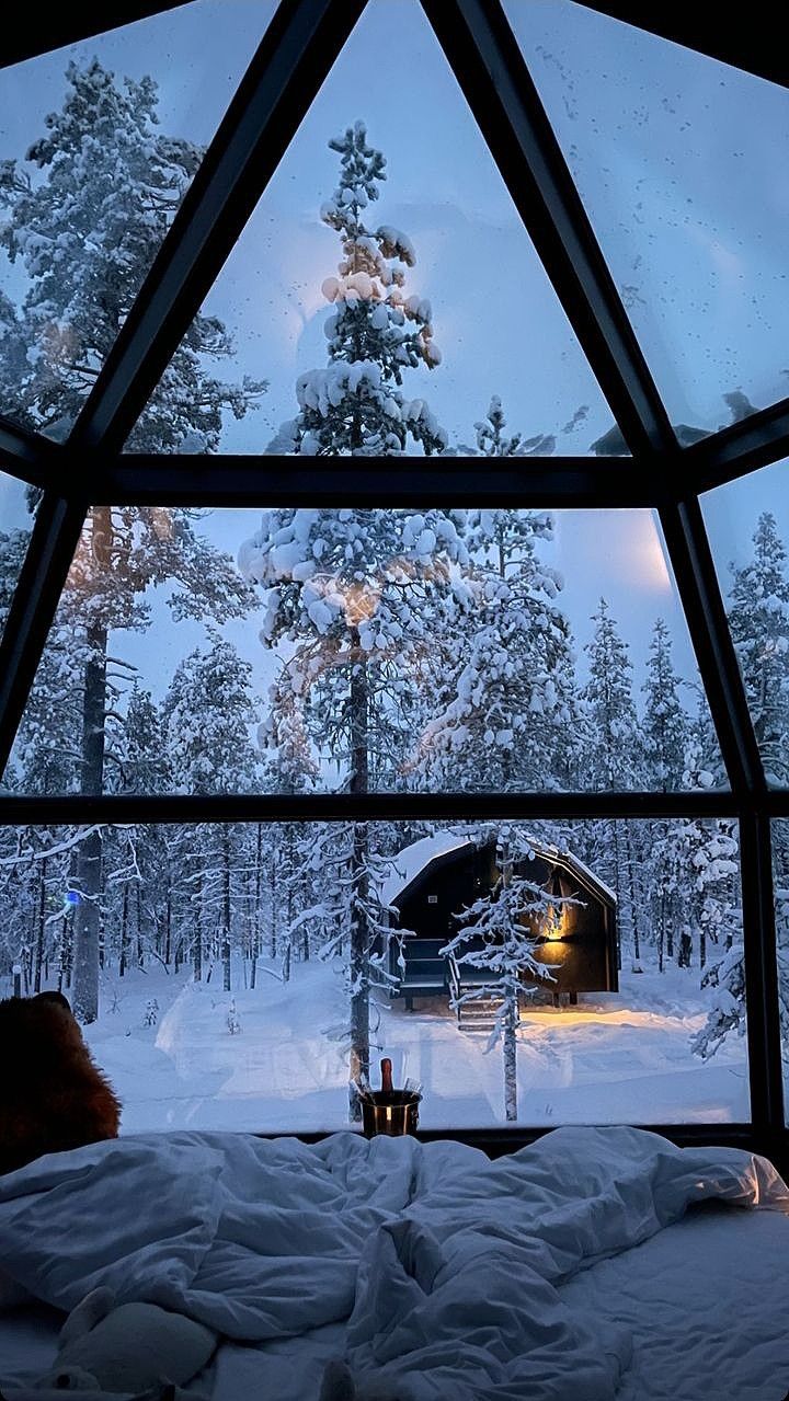 a bedroom with a large window overlooking the snow covered trees and forest at night time