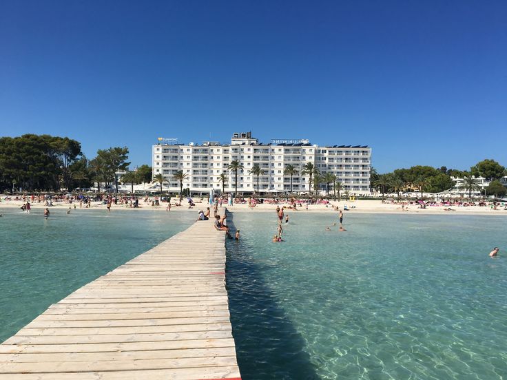 people are swimming in the clear blue water near a beach with white buildings and palm trees