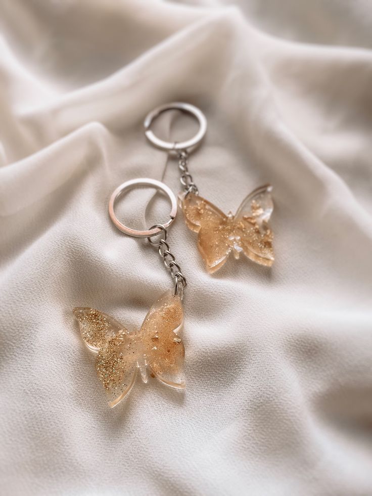 two butterfly shaped key chains on top of a white cloth with gold flecks