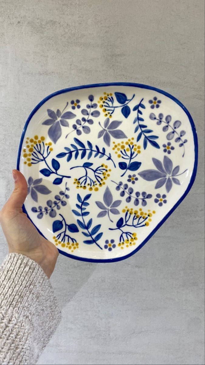 a hand holding a blue and white plate with yellow flowers on the bottom, in front of a gray background