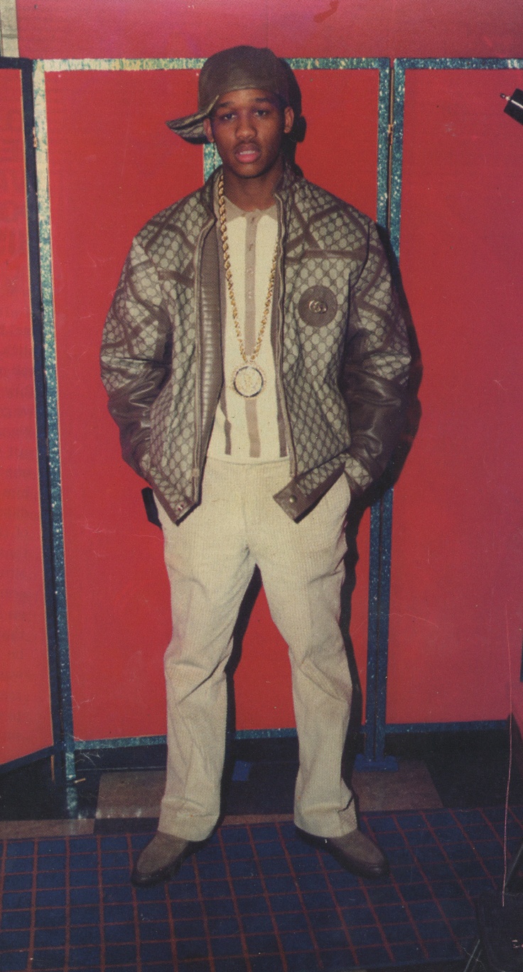 a man standing in front of a red wall wearing white pants and a leather jacket