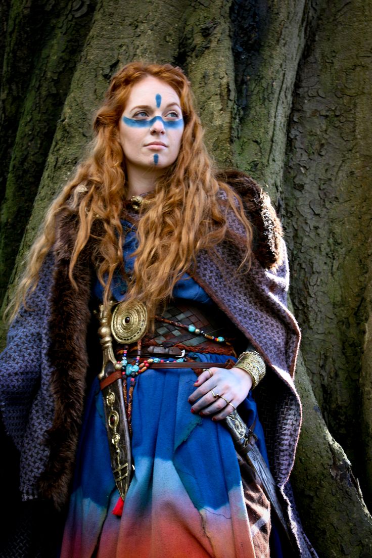 a woman with red hair and blue eyes stands in front of a tree wearing a colorful dress