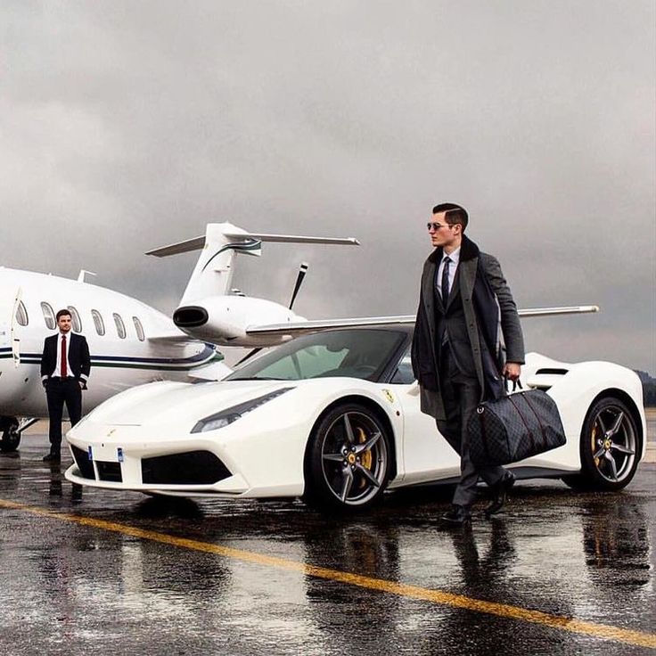 two men in suits standing next to a white sports car with a private jet behind them