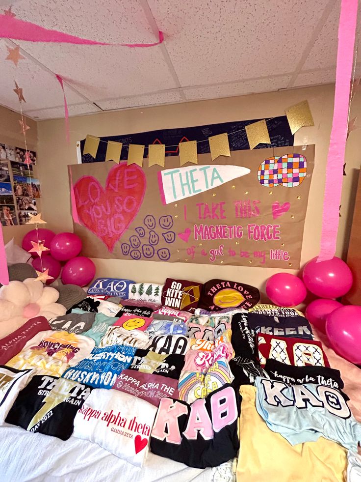 a bed topped with lots of pink and white balloons next to a wall covered in t - shirts