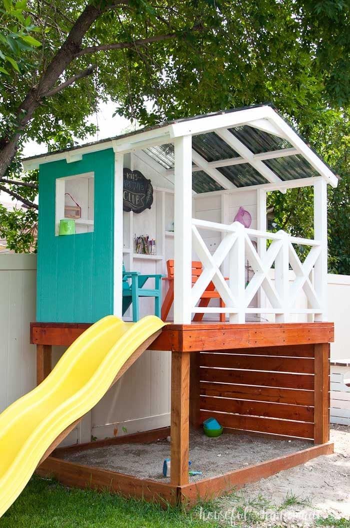 a play house with a slide in the grass next to it and a white fence behind it