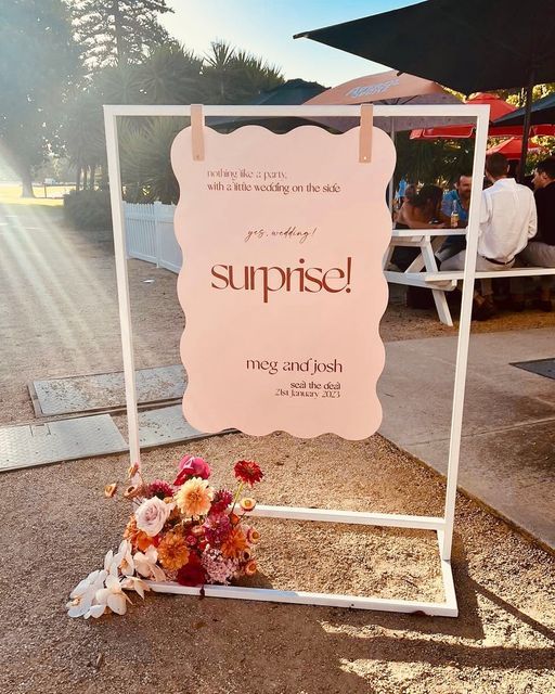 there is a sign that says surprise with flowers on the ground and people in the background