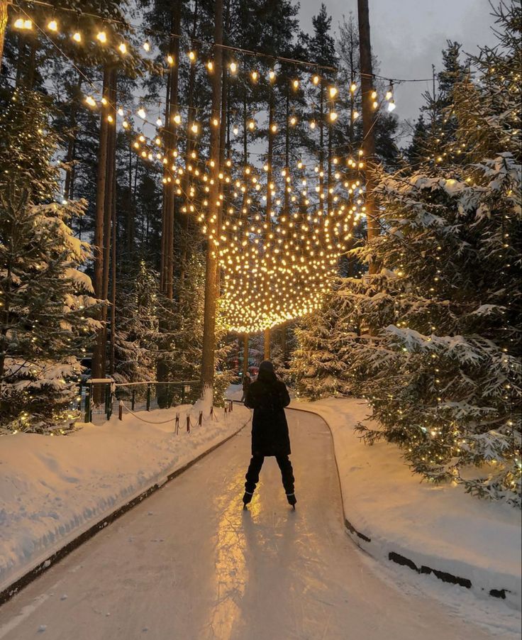 a person standing in the middle of a snow covered road with christmas lights strung over it