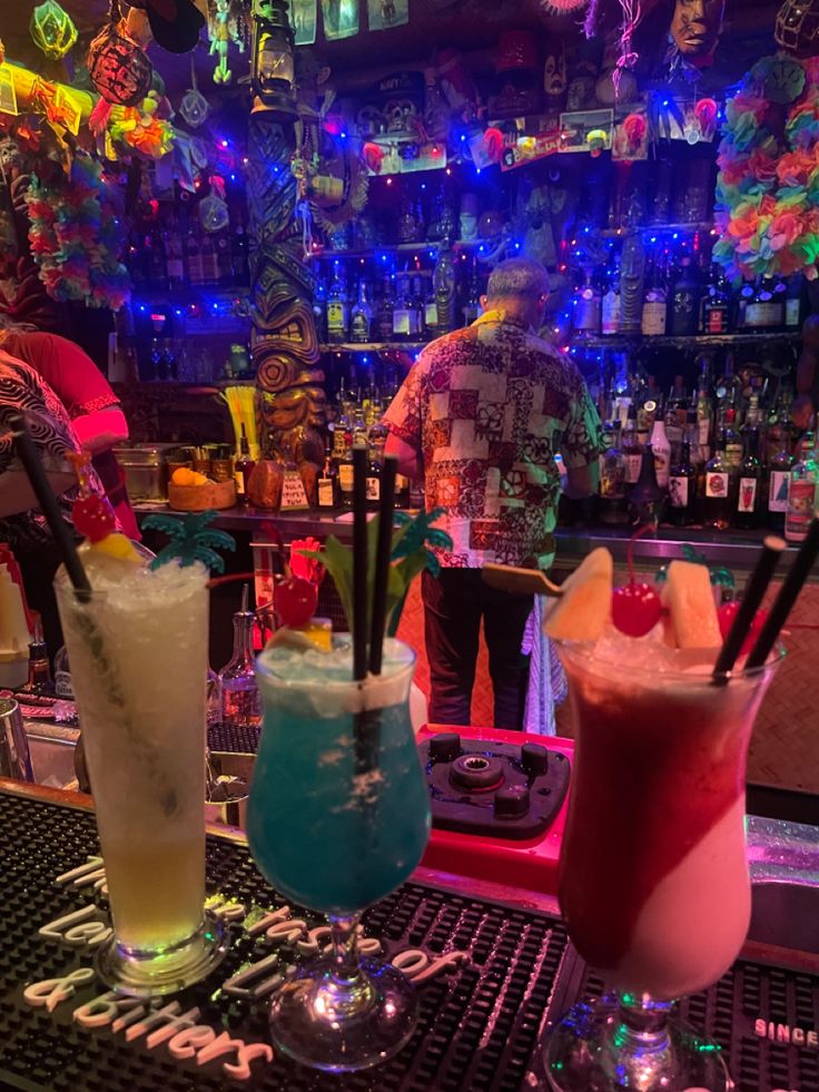 three cocktails are lined up on the bar with colorful lights in the back ground