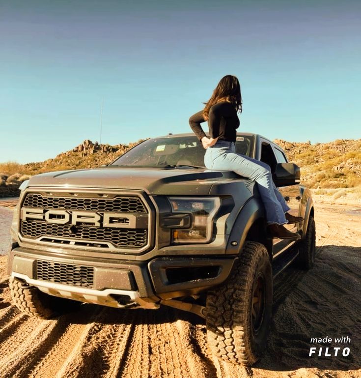 a woman is sitting on the hood of a truck in the desert with her legs crossed