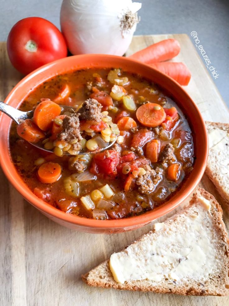 a bowl of soup with carrots, potatoes and meat on a cutting board next to garlic bread