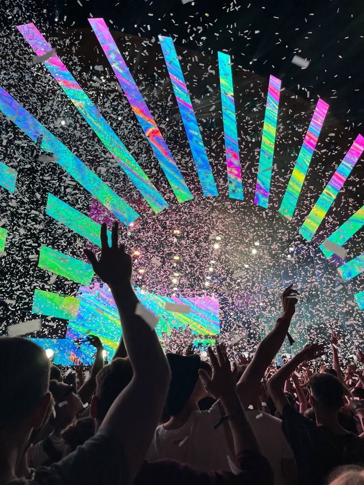 a large crowd at a concert with confetti falling from the sky and people holding their hands in the air