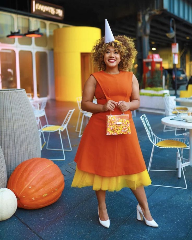 a woman wearing an orange dress and a party hat standing in front of pumpkins