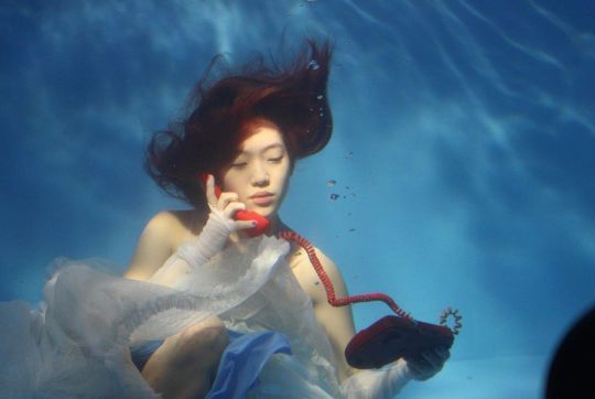 a woman in a white dress is talking on a red phone under the blue water