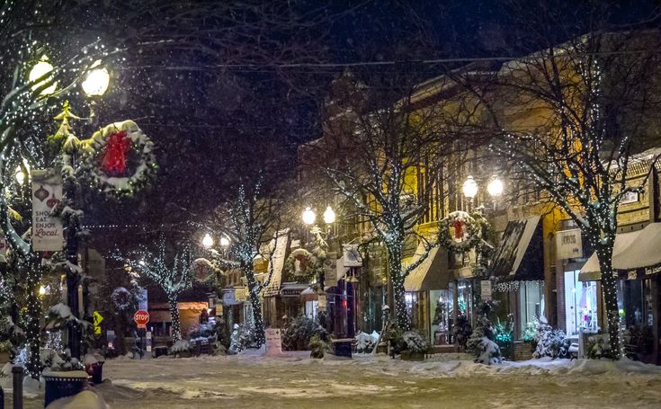 a city street covered in snow at night with christmas decorations on the trees and lights